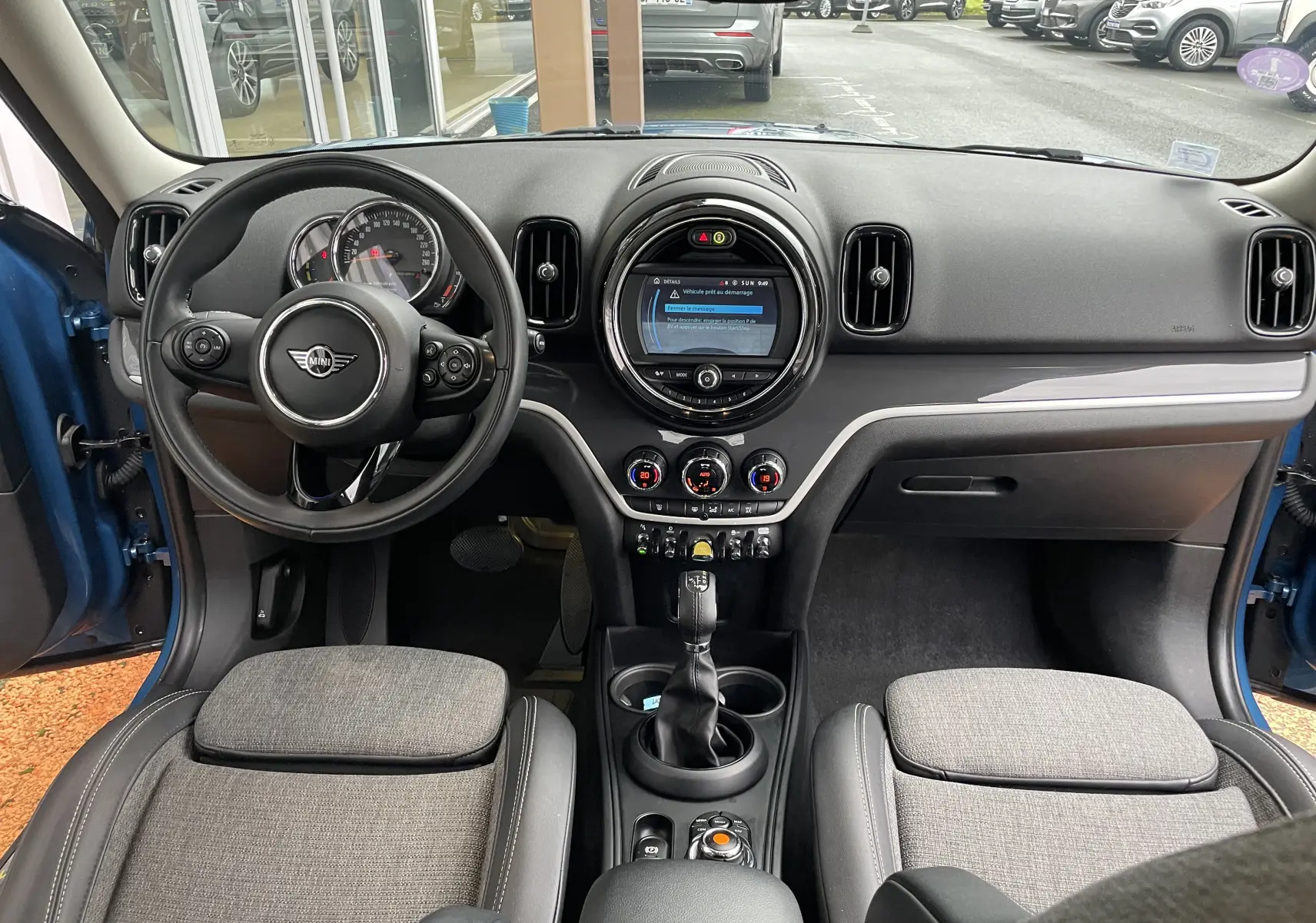Vue intérieure avant du MINI Countryman Cooper S hybride rechargeable, sièges gris et tableau de bord noir avec écran central rond.