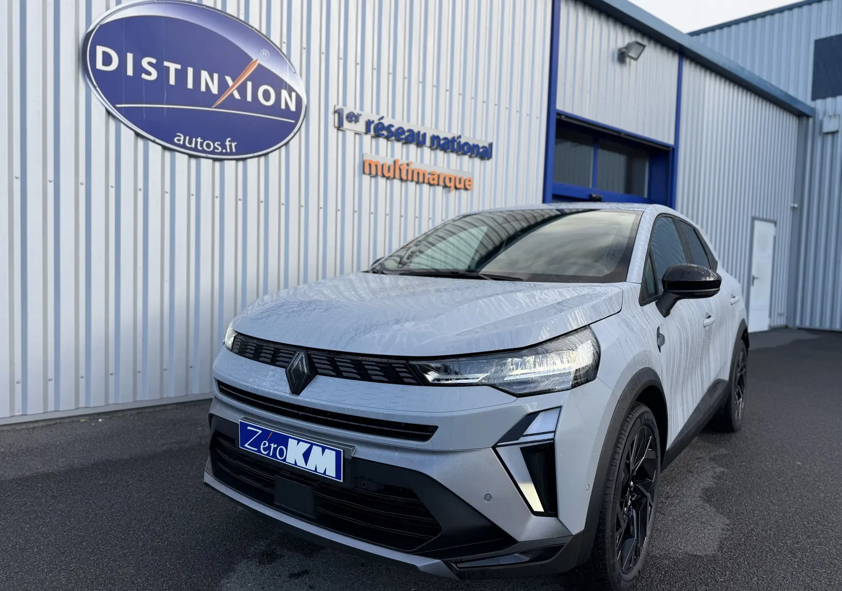 Vue 3/4 avant d'une Renault Symbioz gris rafale avec calandre noire et jantes noires devant un bâtiment industriel.