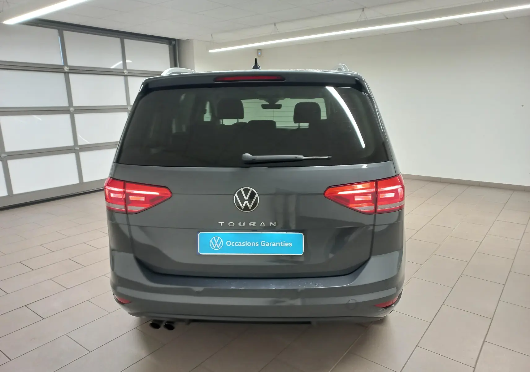 Vue arrière d'un Volkswagen Touran 2025 gris dauphin avec feux allumés dans un garage lumineux.
