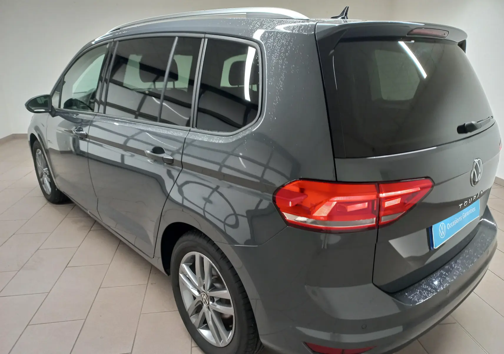 Vue 3/4 arrière droite d’un Volkswagen Touran 2025 gris dauphin métallisé avec feux arrière allumés en intérieur.