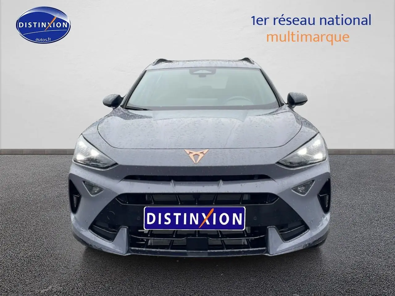Vue avant d'un CUPRA Formentor gris graphene 2026 avec logo cuivré et phares LED allumés sur sol humide.
