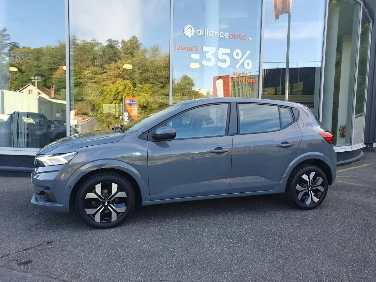 Profil côté gauche d'une Dacia Sandero 2024 gris urbain avec jantes alu 16 pouces, garée devant une vitrine.