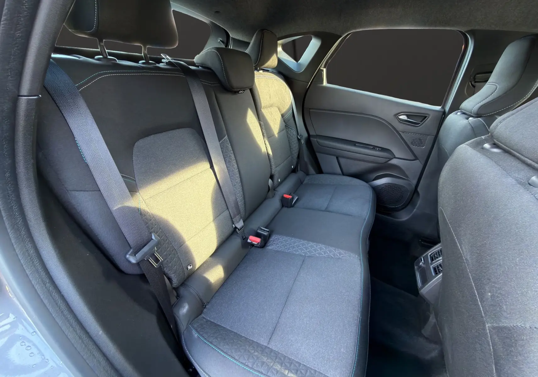 Vue intérieure arrière côté gauche du Renault Captur 2024, sièges tissu gris avec surpiqûres bleues et ceintures de sécurité.