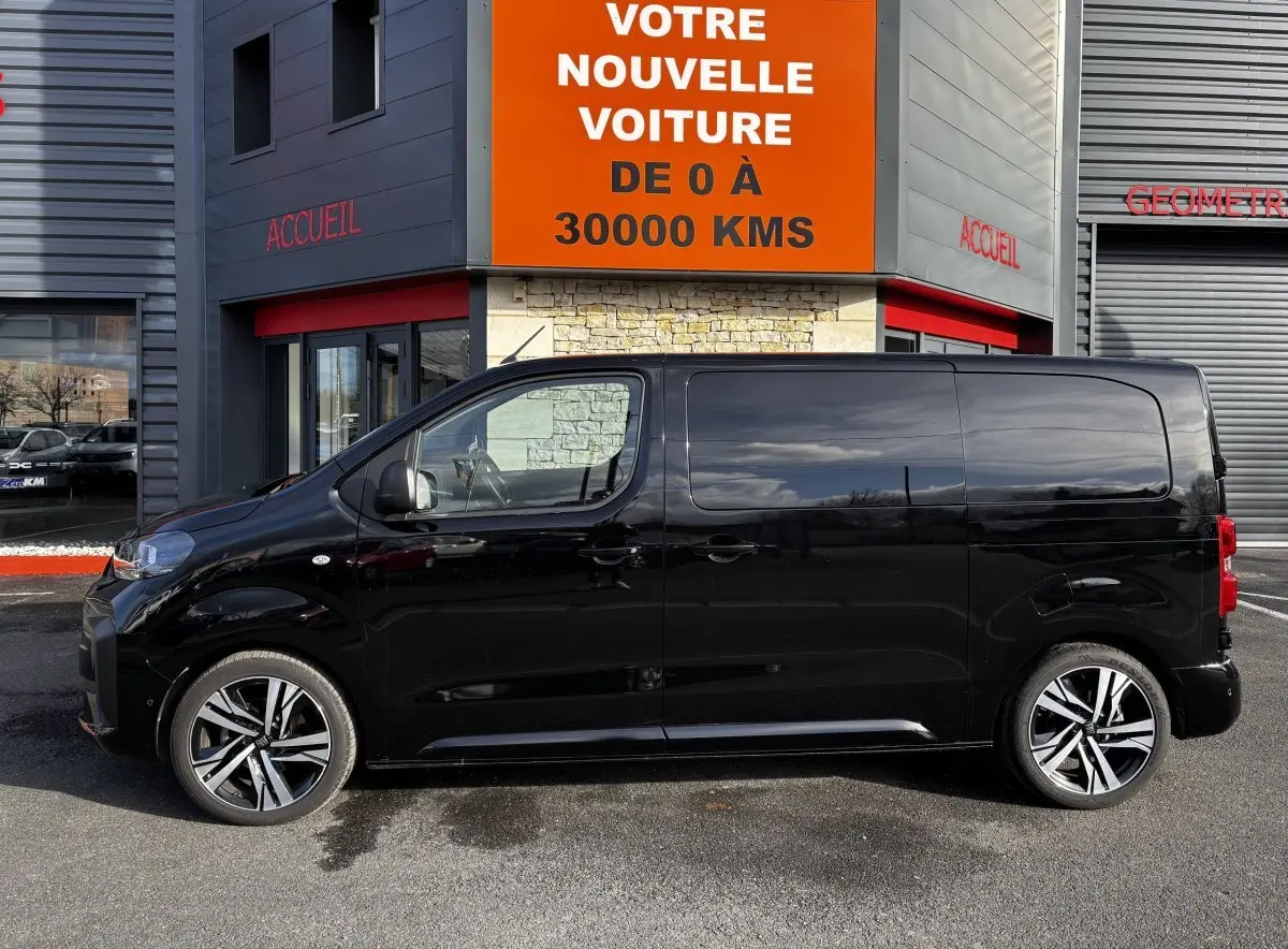 Profil côté gauche d’un FIAT Scudo Fourgon noir 2025 garé devant un bâtiment avec enseigne orange.