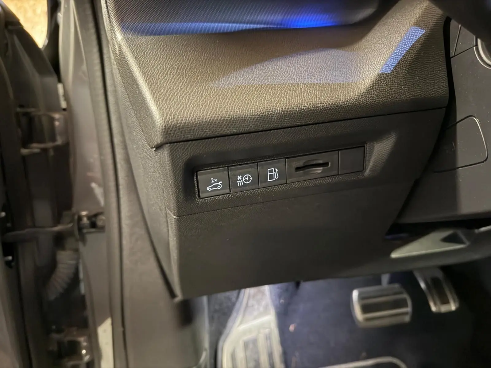 Vue intérieure côté conducteur du tableau de bord noir avec commandes d'ouverture de coffre et réglages, pédales aluminium visibles.