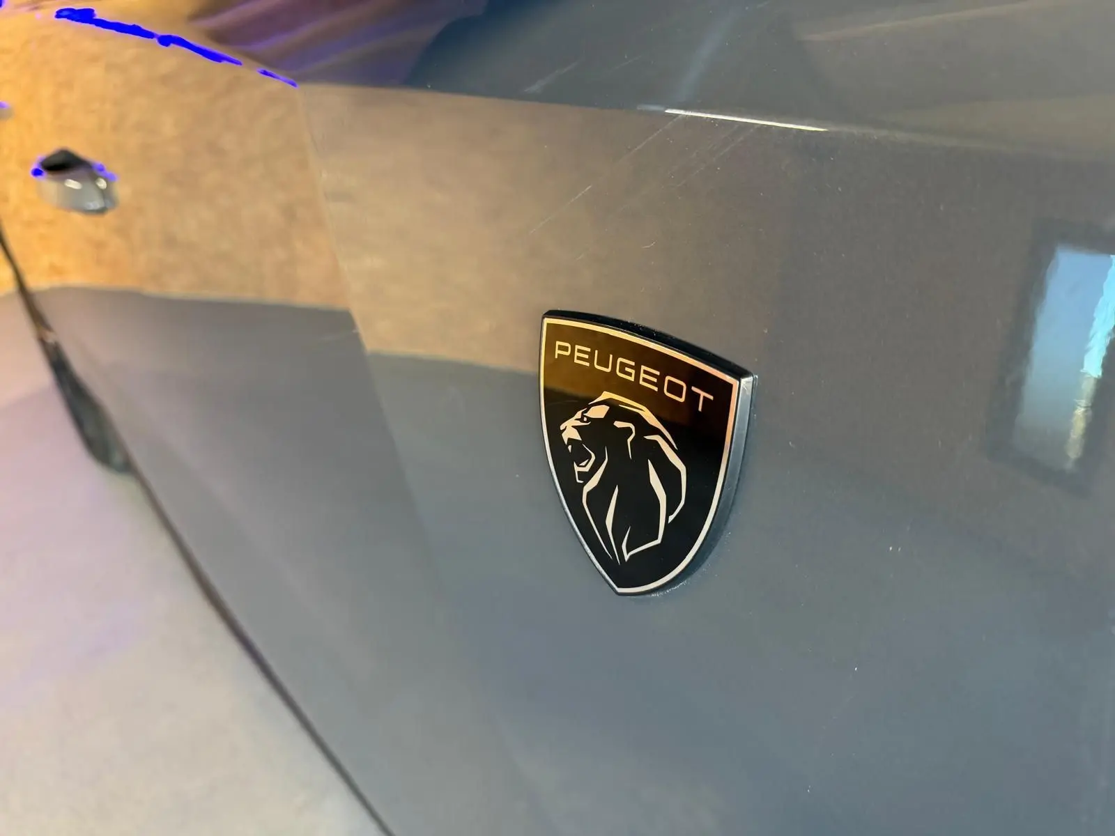 Gros plan sur l’écusson Peugeot noir brillant sur la porte côté gauche d’une 408 gris métallisé.