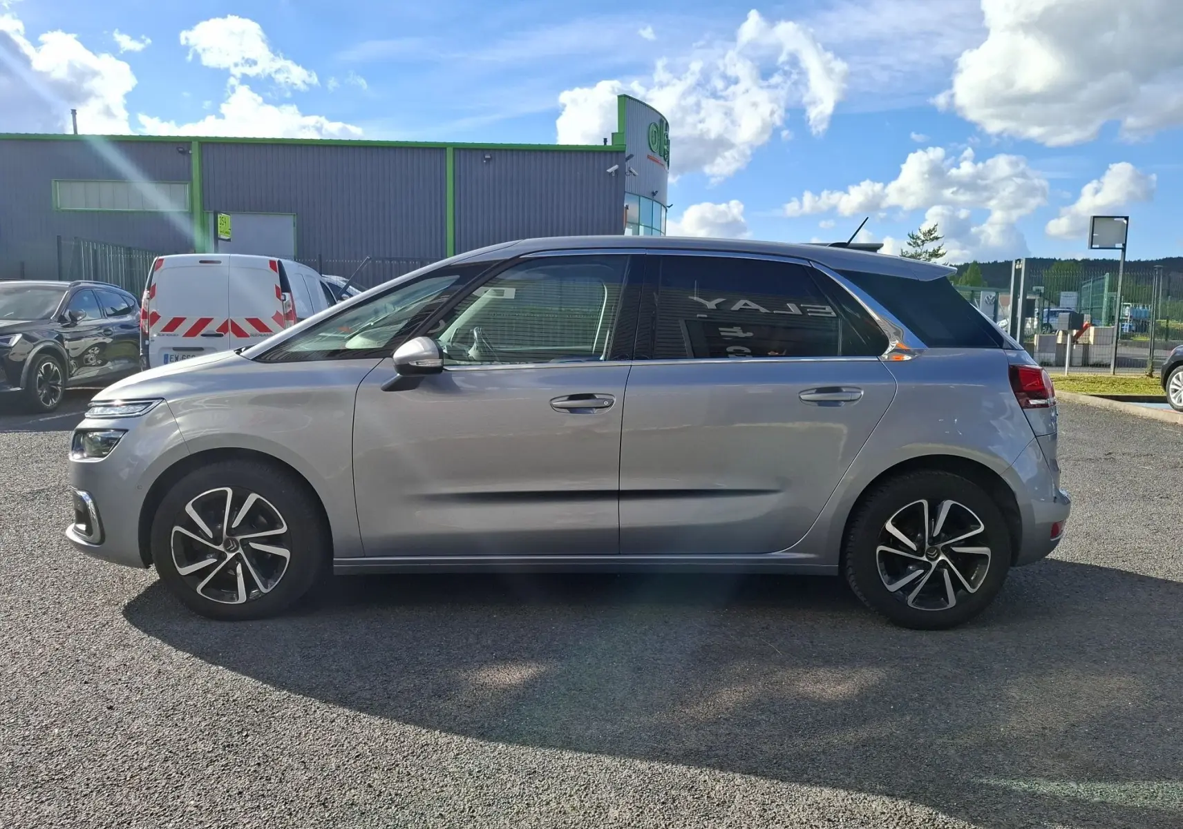 Vue de profil côté gauche d'une Citroën C4 Picasso gris clair avec jantes alliage 17 pouces sur parking extérieur.
