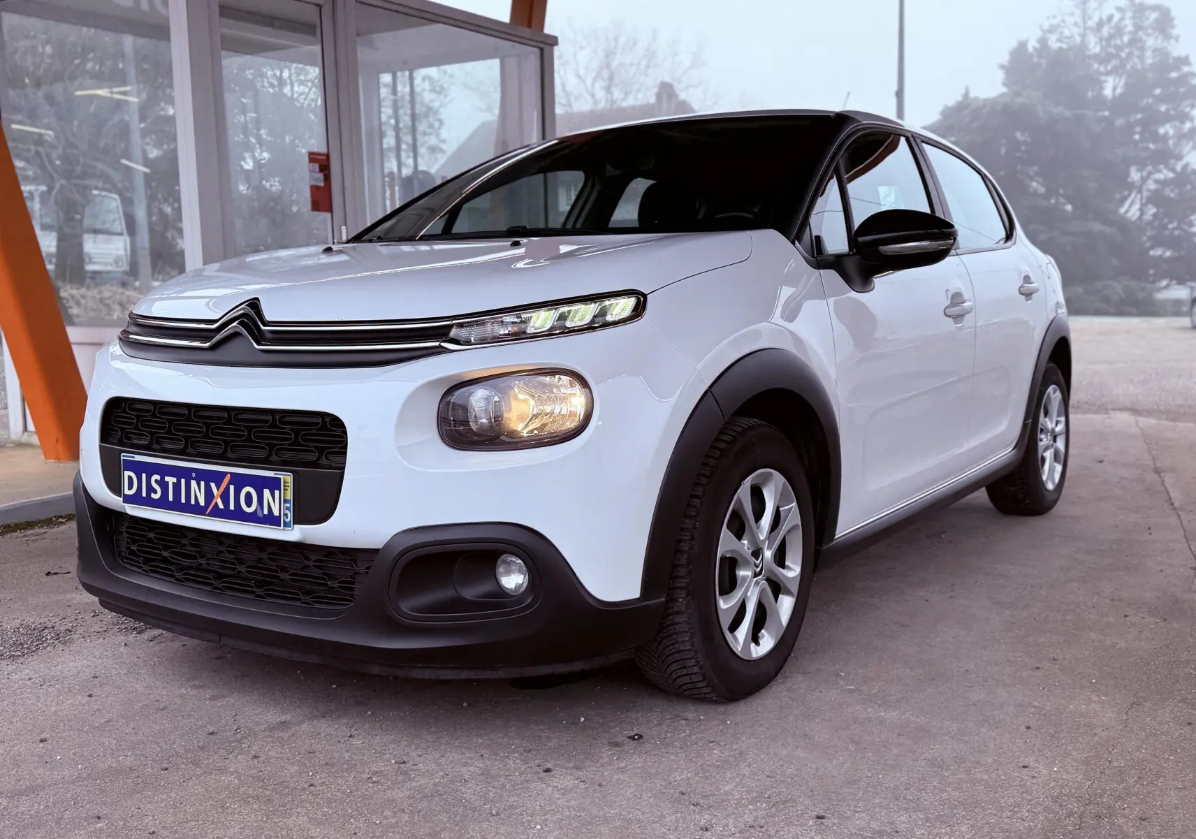 Citroën C3 blanc vue 3/4 avant droit, avec calandre noire et phares allumés sous abri.