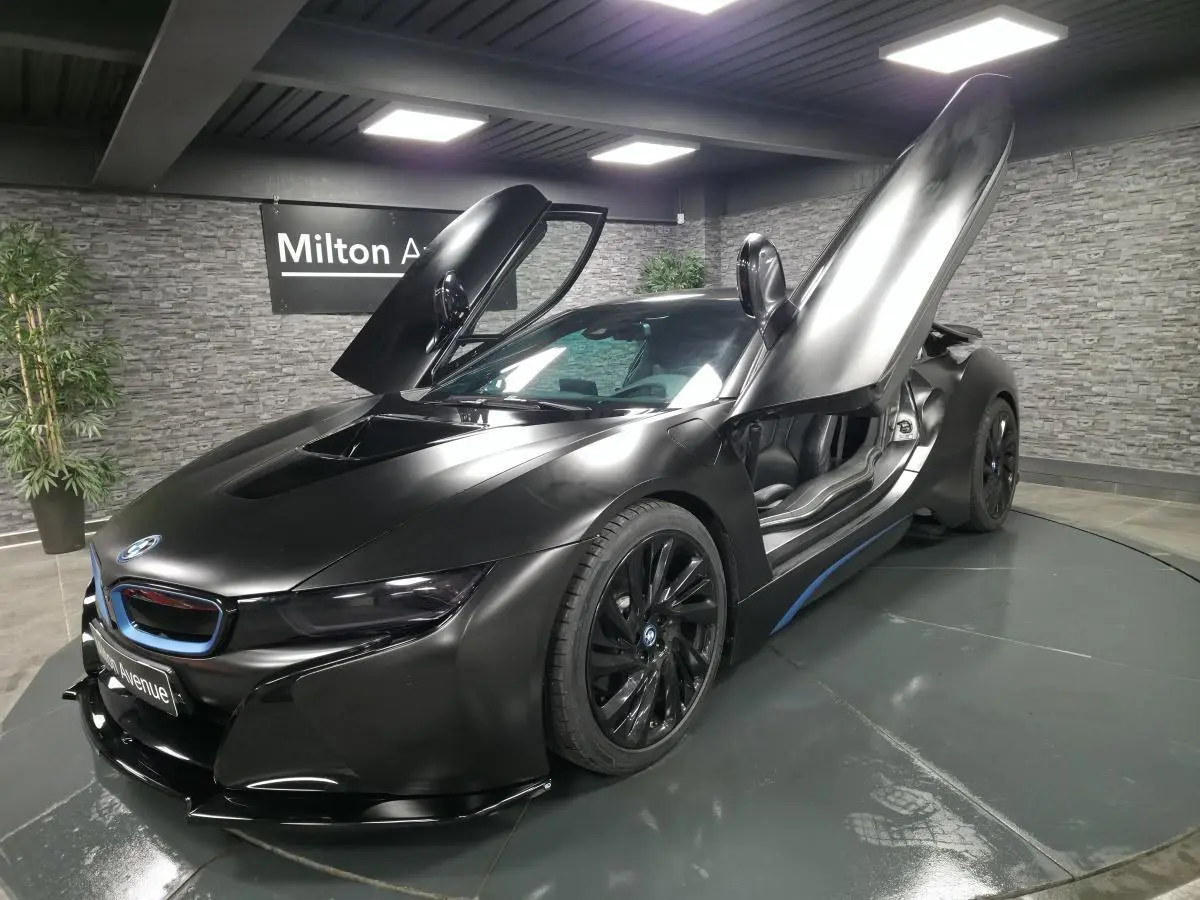 BMW i8 Coupé Pure Impulse noir mat vue 3/4 avant droit avec portes en élytre ouvertes dans un showroom.