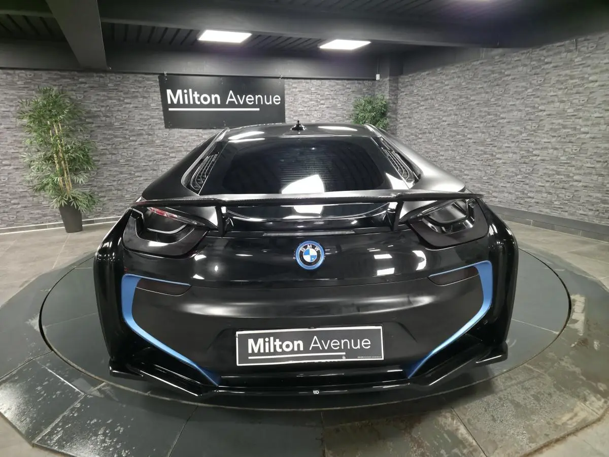 Vue arrière d'une BMW i8 Coupé Pure Impulse noir mat avec détails bleus et aileron carbone en intérieur showroom.