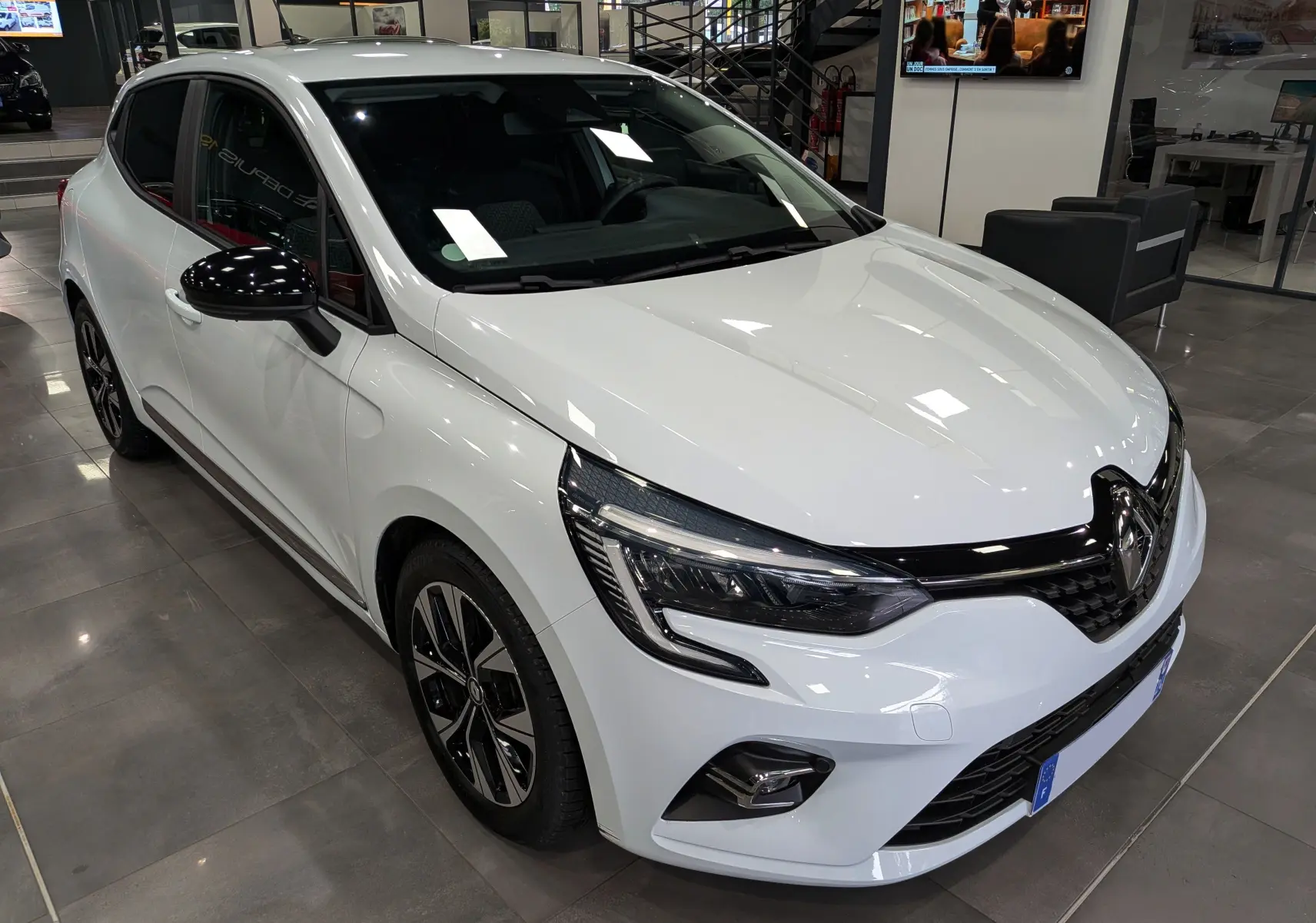Renault Clio V blanc glacier vue 3/4 avant droit, avec jantes noires et rétroviseurs noirs brillants en intérieur showroom.