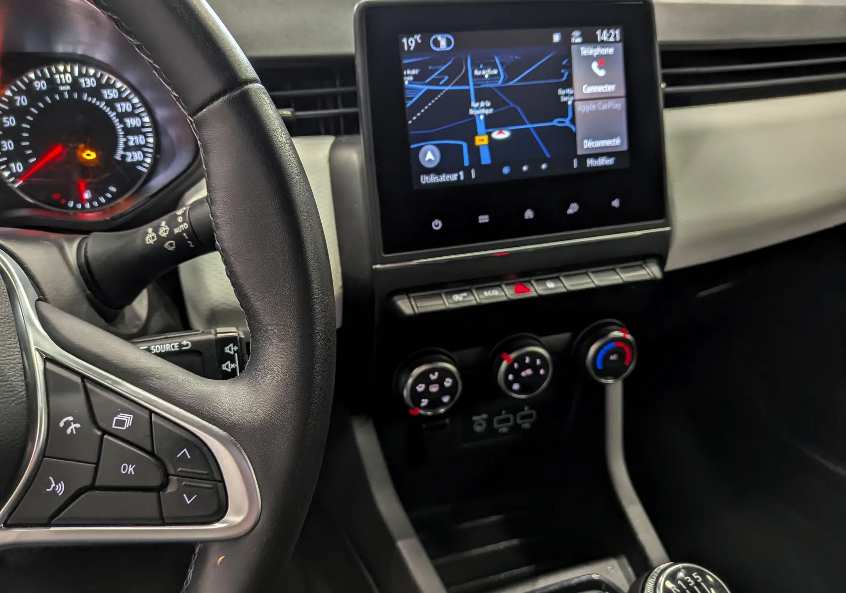 Vue rapprochée de l’intérieur de la Renault Clio V blanc glacier, montrant le volant, l’écran tactile avec GPS et la boîte manuelle.