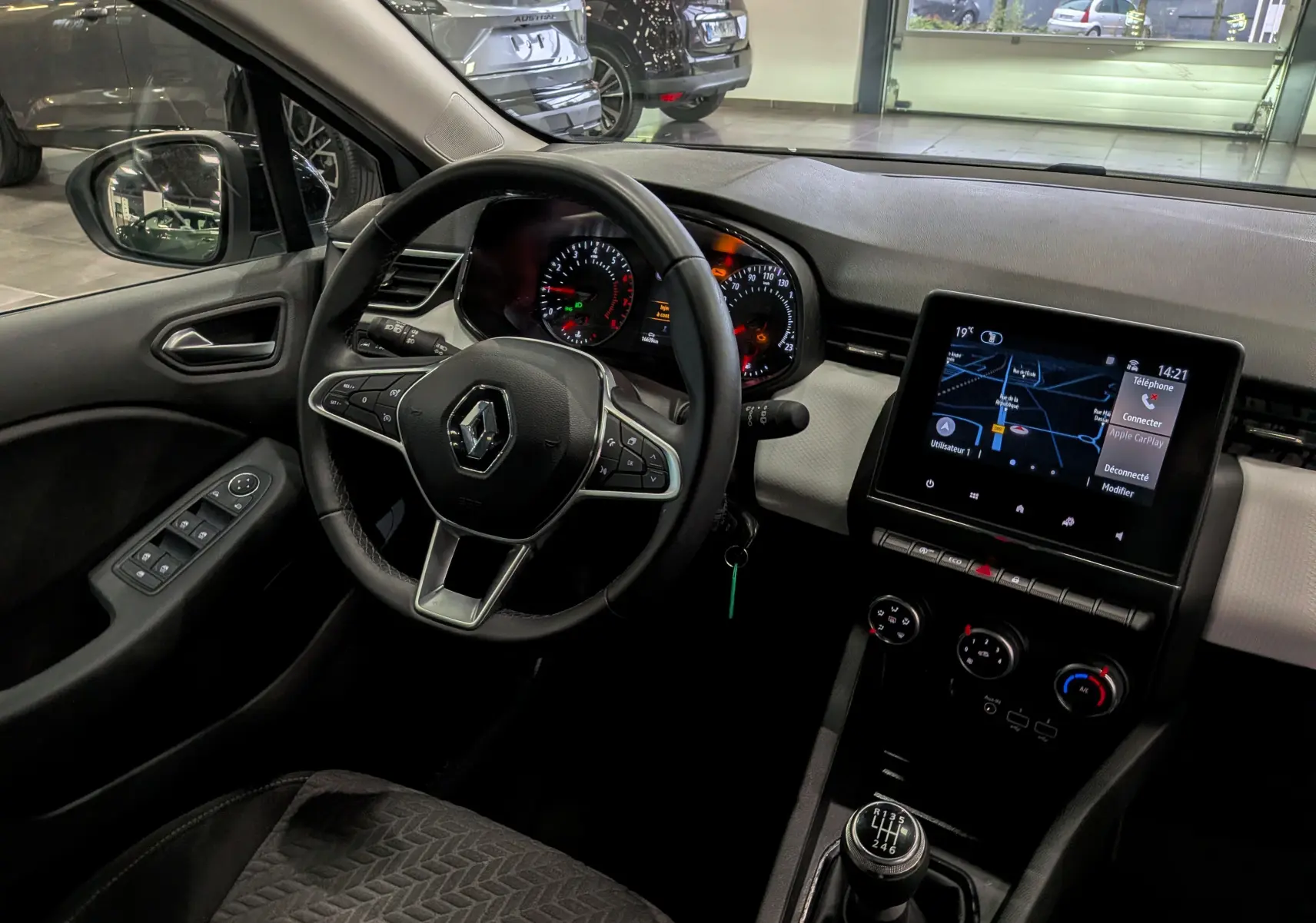 Vue intérieure côté conducteur de la Renault Clio V blanc glacier, avec tableau de bord, volant multifonction et écran tactile central.