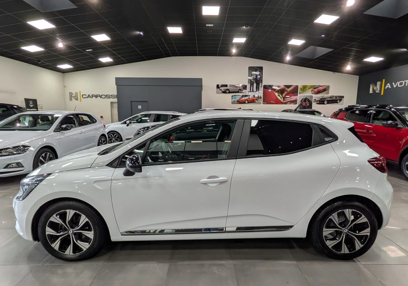 Profil latéral droit d'une Renault Clio V blanche Glacier, avec jantes alliage noires et détails chromés, en showroom.