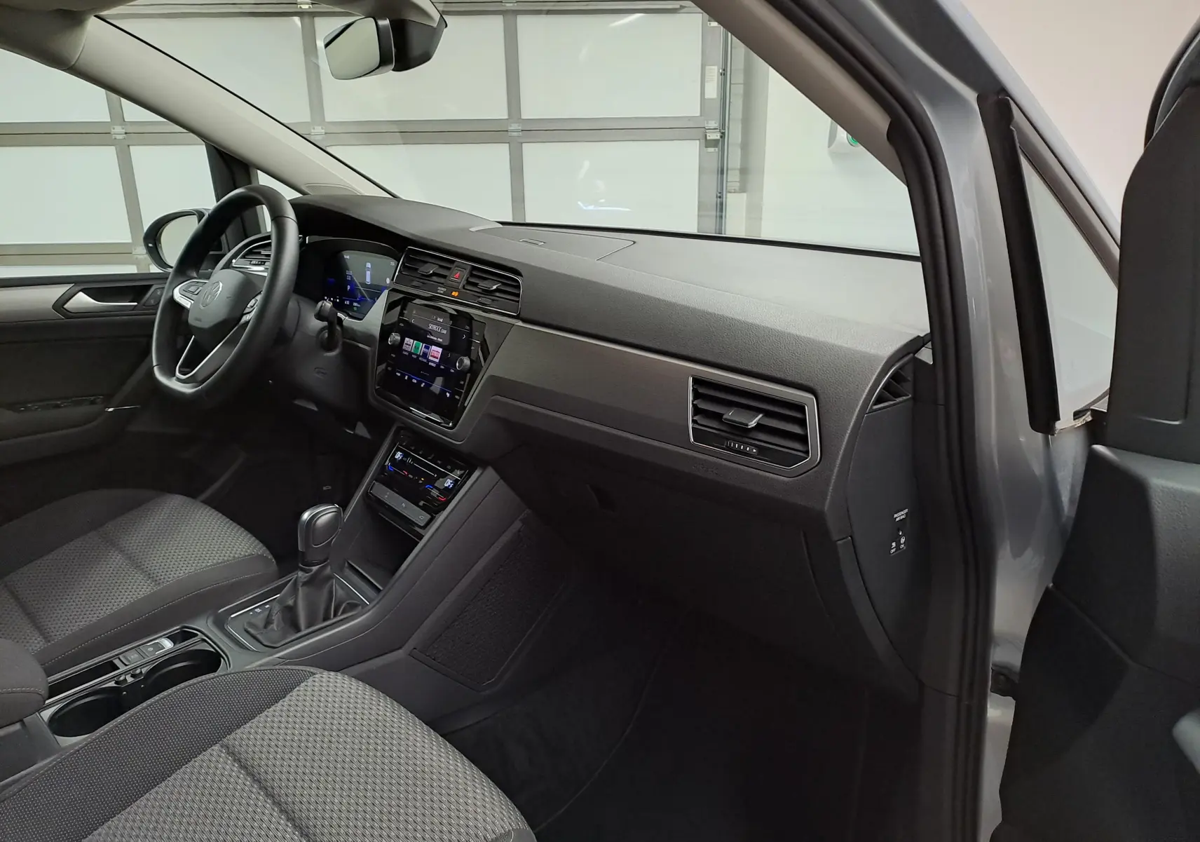 Vue intérieure côté passager du Volkswagen Touran 2025 argent Dolomite, montrant tableau de bord et sièges en tissu gris.
