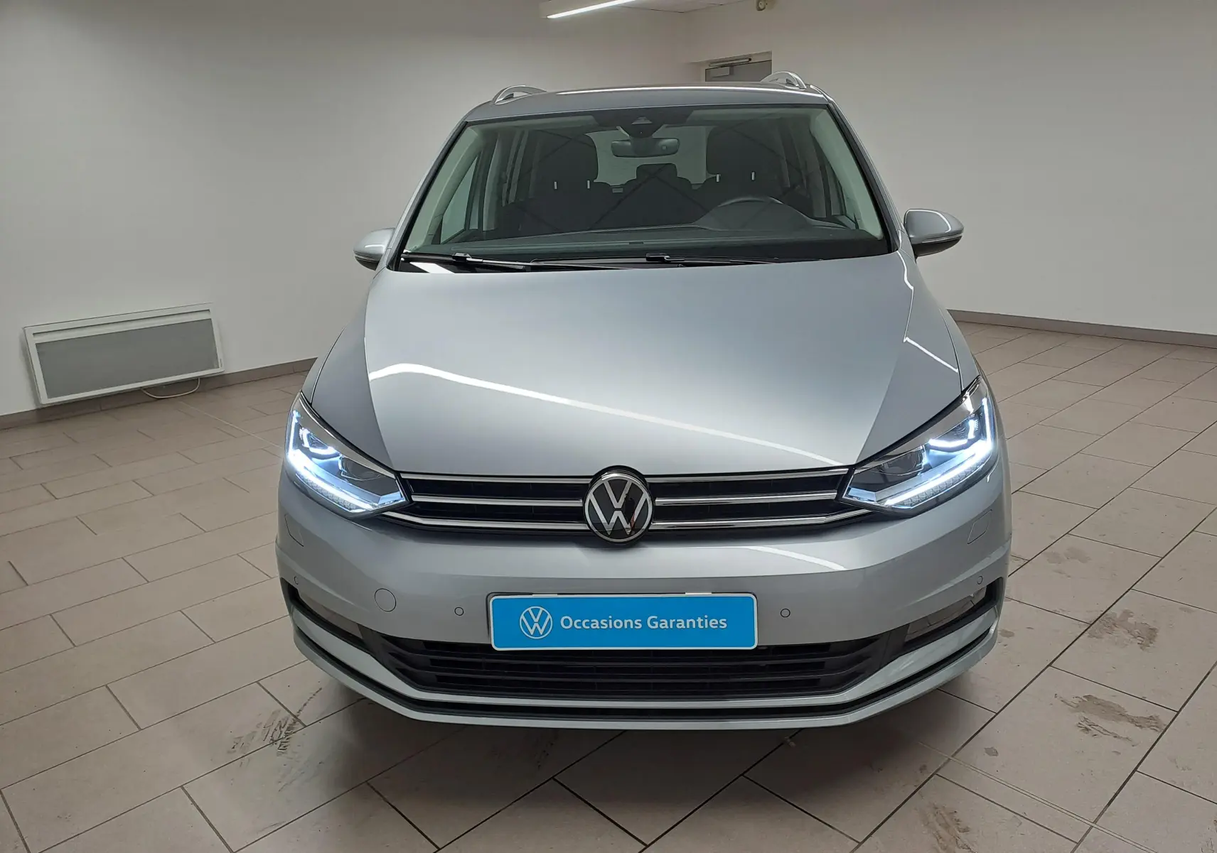 Vue frontale d'un Volkswagen Touran 2025 argent Dolomite avec phares allumés dans un showroom.
