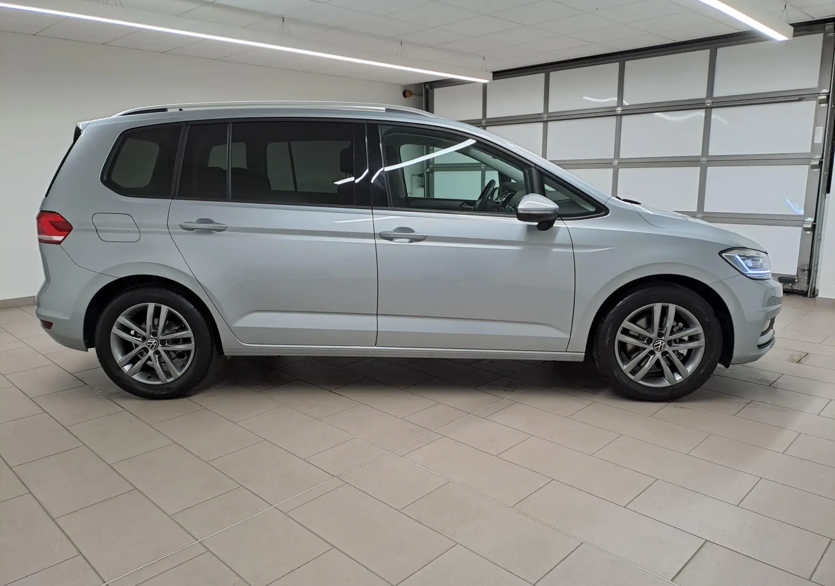 Profil droit d'un Volkswagen Touran 2025 argent Dolomite avec jantes alliage et vitres teintées, en intérieur showroom.