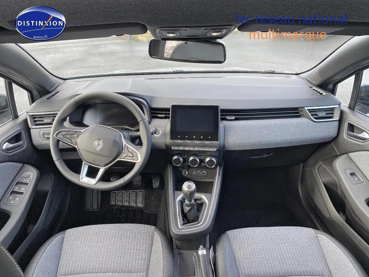 Vue intérieure avant de la Renault Clio V gris rafale, volant multifonction, écran tactile et levier de vitesse manuel visibles.