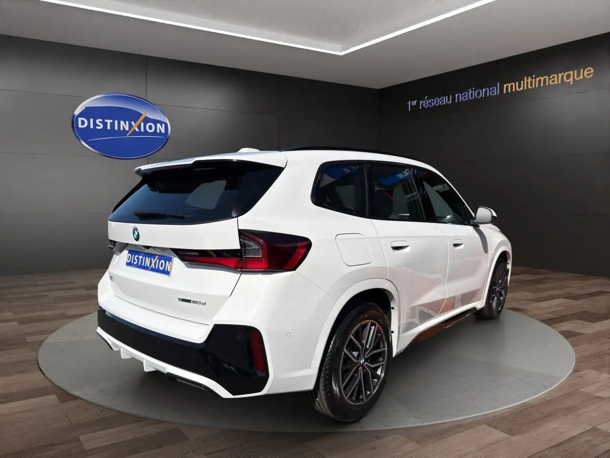 BMW X1 sDrive20d blanc en 3/4 arrière droit avec toit noir et jantes sport dans showroom