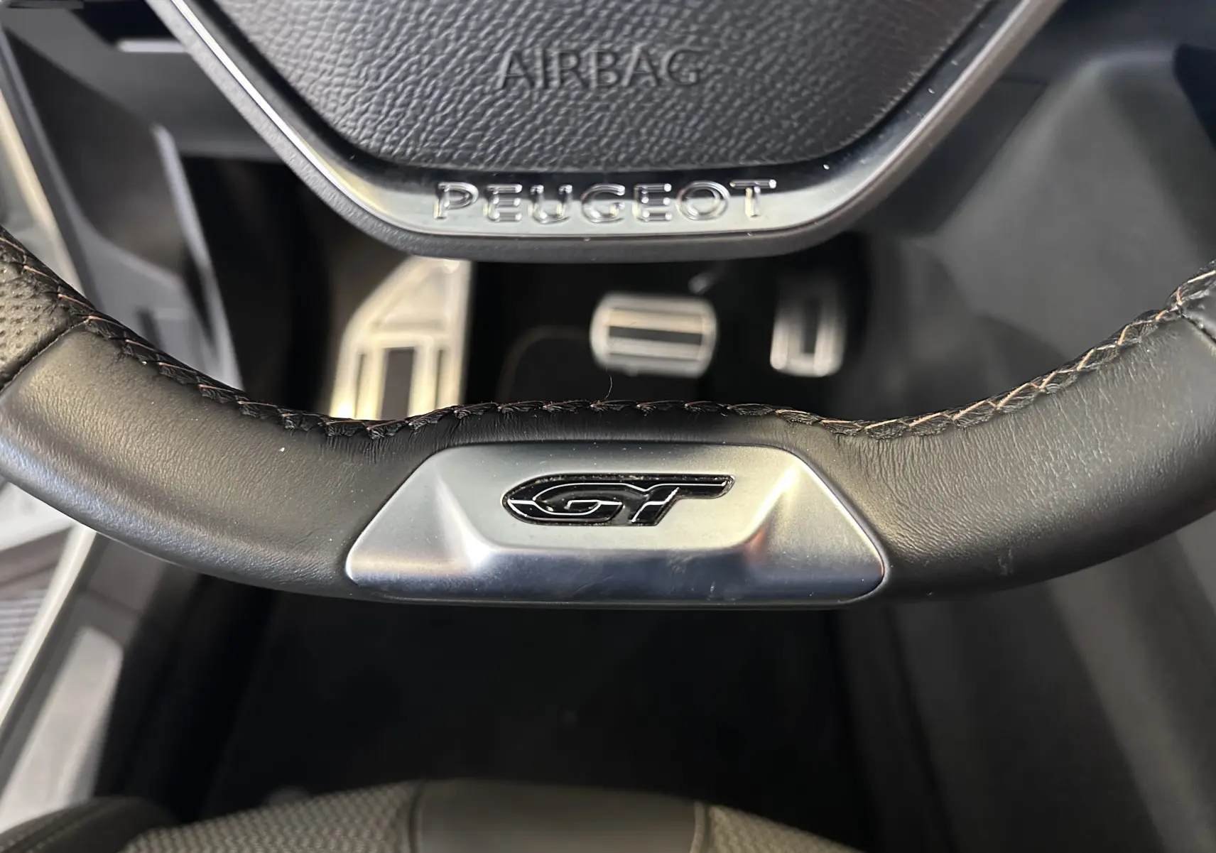 Gros plan sur le volant cuir noir du Peugeot 3008 GT avec logo GT et pédalier en aluminium visible en arrière-plan.