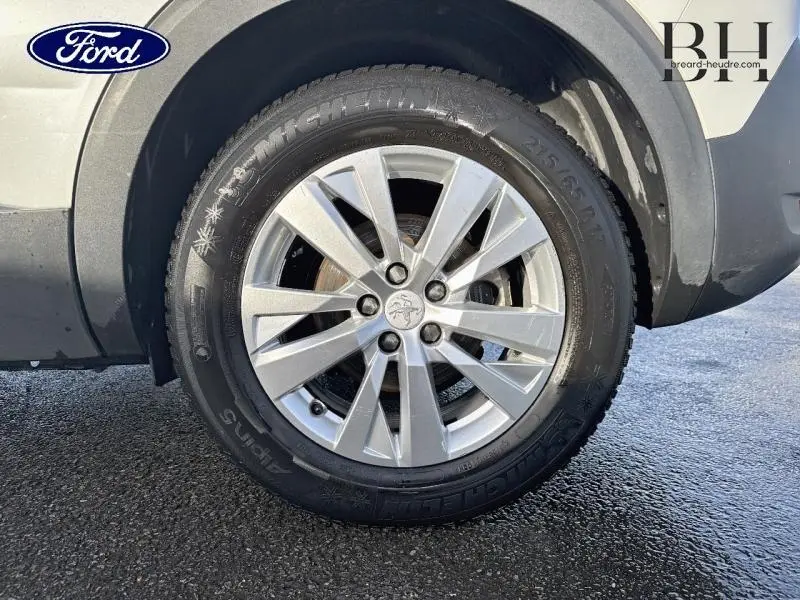 Gros plan sur la roue avant droite d'un Peugeot 5008 blanc Banquise, avec jante alu et logo Peugeot central.