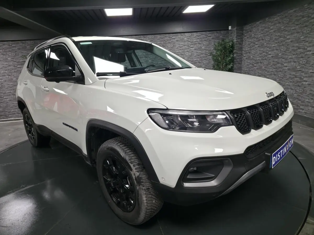 Jeep Compass blanc vue 3/4 avant droit avec jantes noires et calandre distinctive en intérieur showroom.