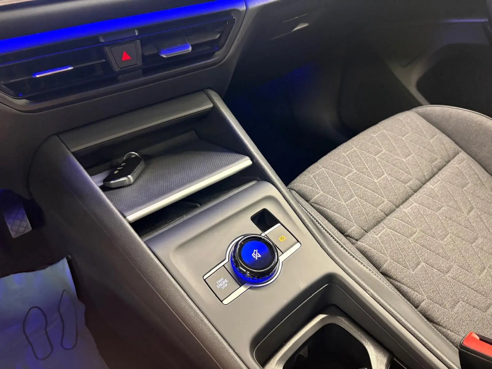Gros plan sur la console centrale du Volkswagen Tiguan 2025, avec molette de commande éclairée en bleu et siège tissu gris.