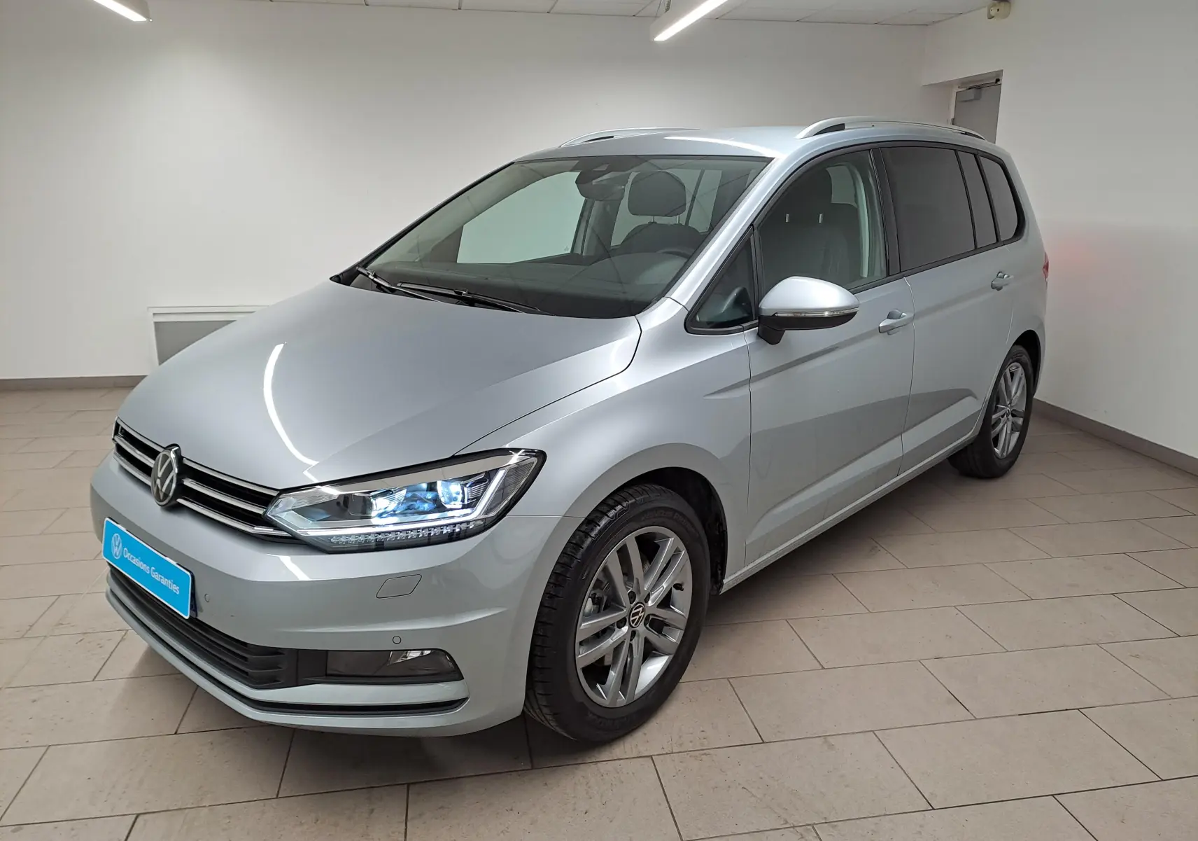 Volkswagen Touran 2025 argent Dolomite en 3/4 avant droit, avec phares LED allumés et jantes alliage.