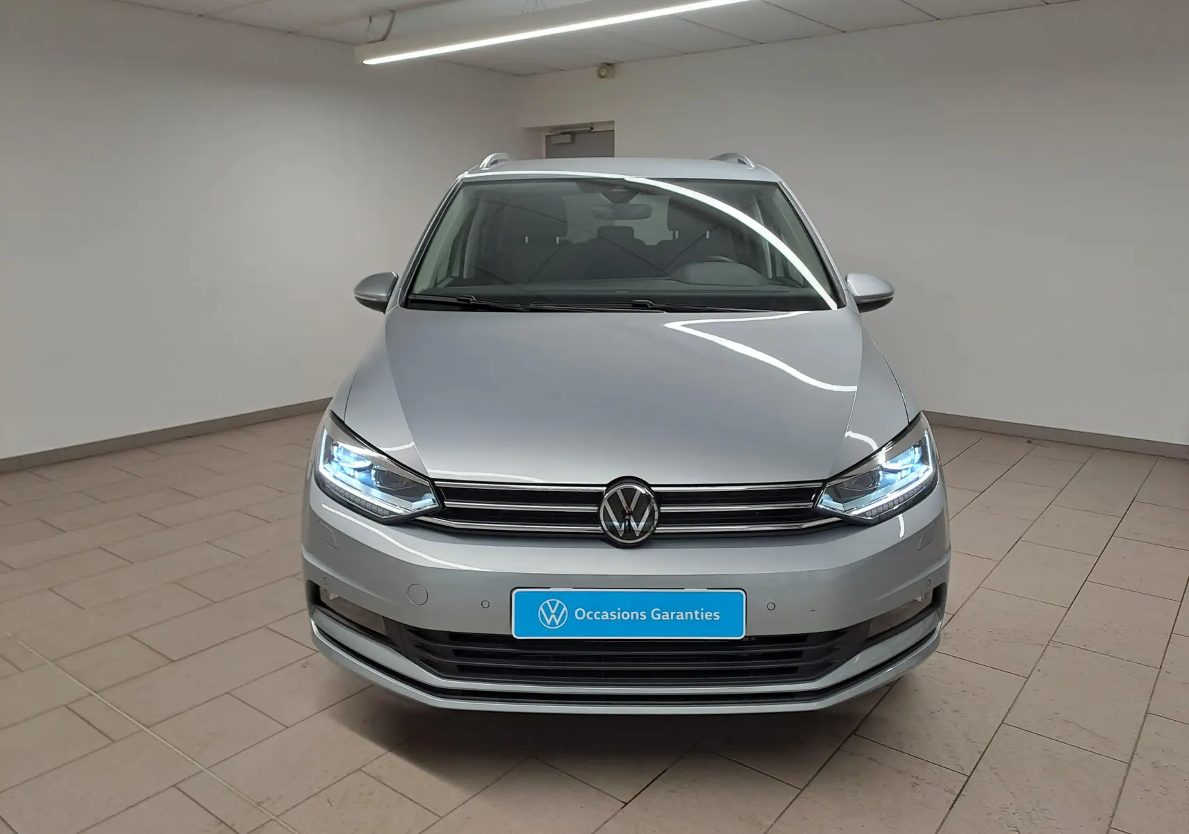 Vue avant frontale du Volkswagen Touran 2025 en argent Dolomite avec phares allumés et plaque "Occasions Garantis" visible.