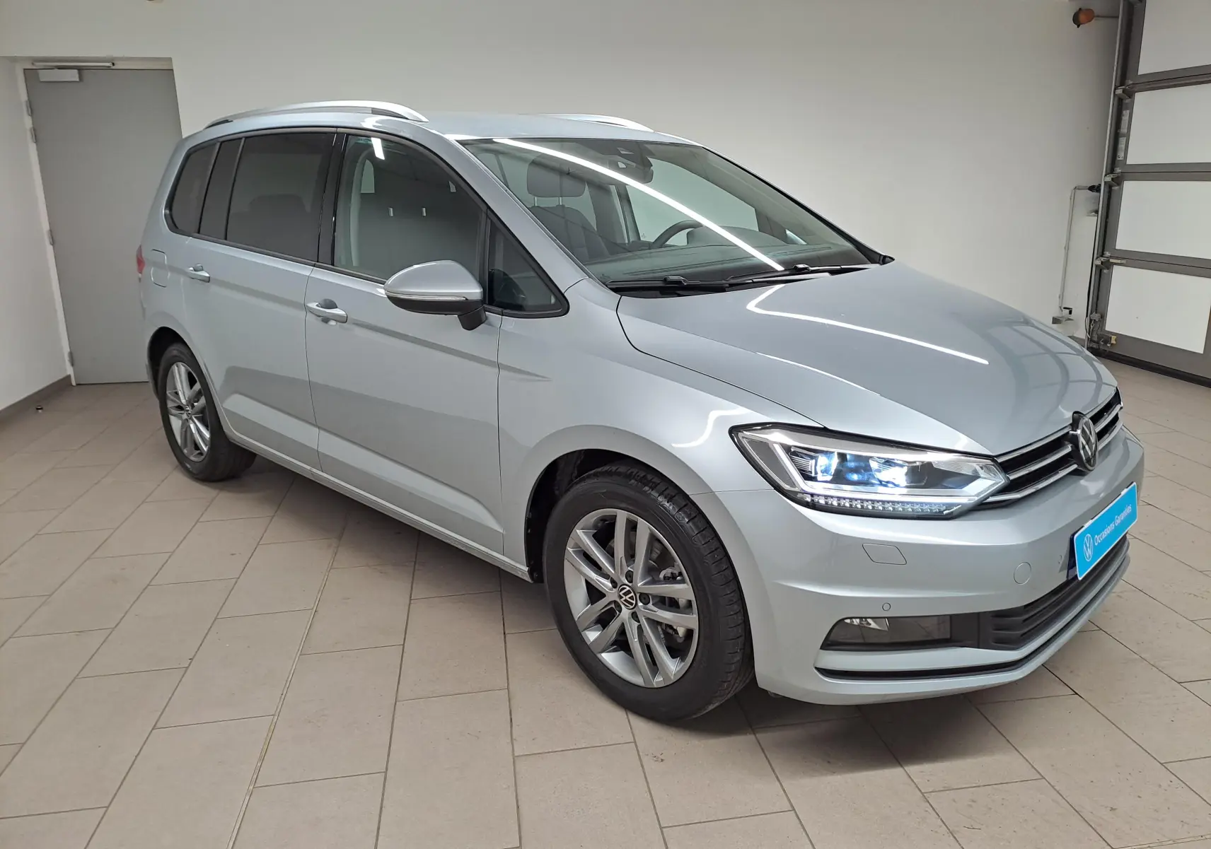 Volkswagen Touran 2025 argent Dolomite en 3/4 avant droit, avec phares allumés et jantes alliage visibles.