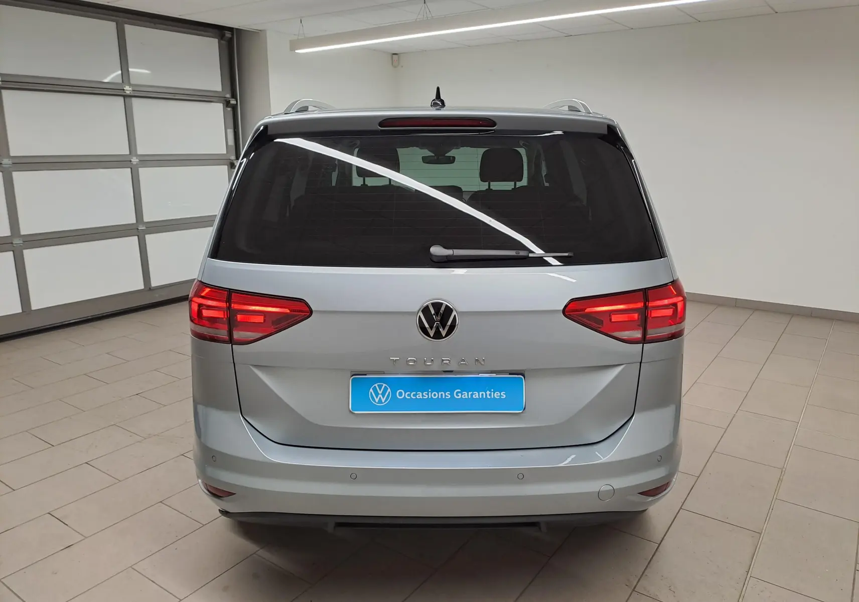 Vue arrière d'un Volkswagen Touran 2025 argent Dolomite avec feux allumés et plaque "Occasions Garantis" en intérieur.