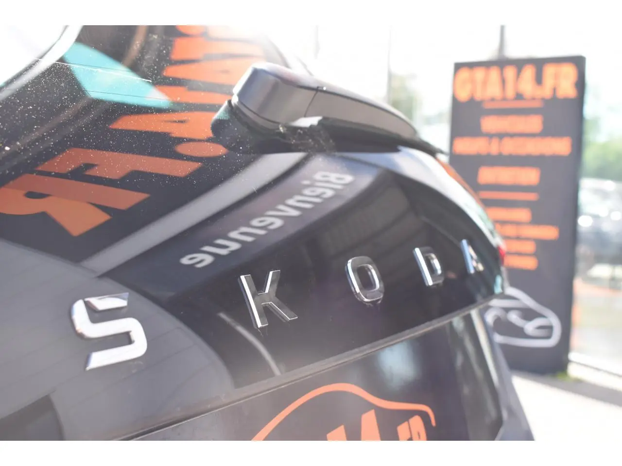 Gros plan sur l’arrière gris graphite d’une Skoda Scala 2025 avec logo SKODA en relief et reflet d’enseigne orange.