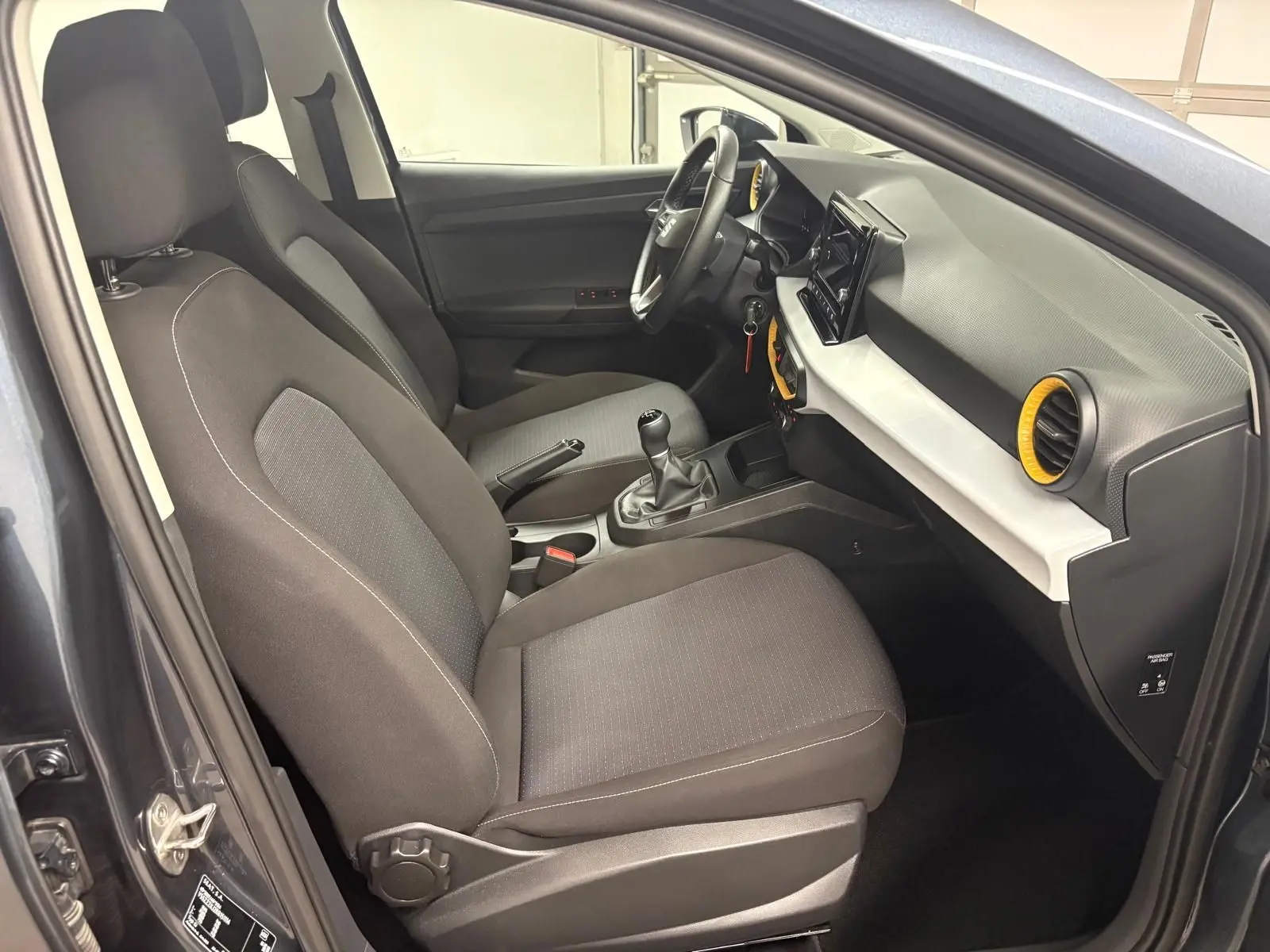 Intérieur avant droit de la SEAT Ibiza 2024 gris magnétique, avec volant, boîte manuelle et aérateurs jaunes distinctifs.