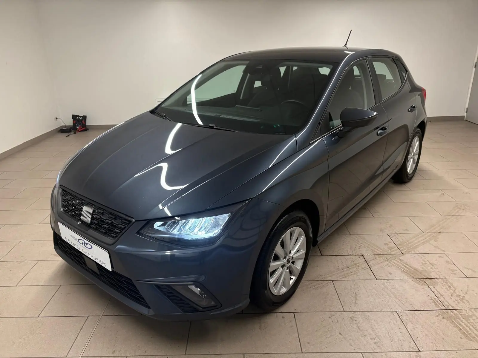 SEAT Ibiza 2024 gris magnétique vue 3/4 avant droit avec phares allumés dans un intérieur neutre.