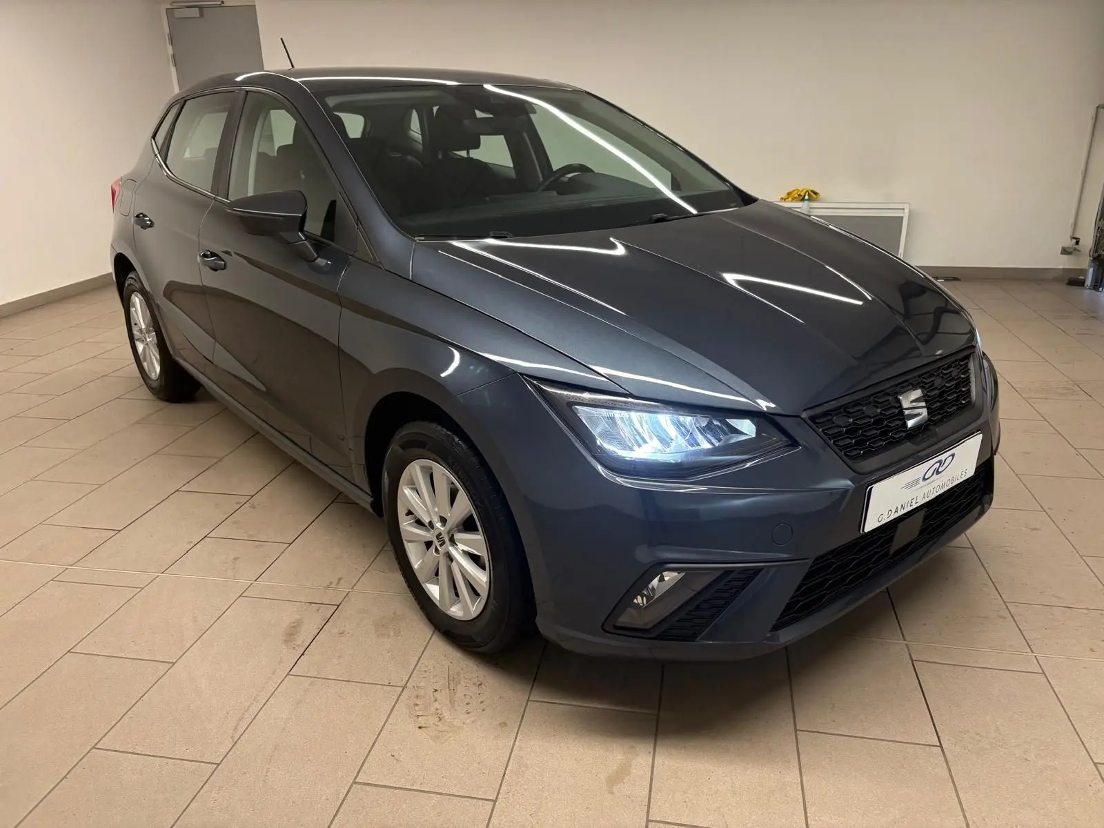 Vue 3/4 avant d'une SEAT Ibiza gris magnétique 2024 avec phares allumés dans un garage intérieur.