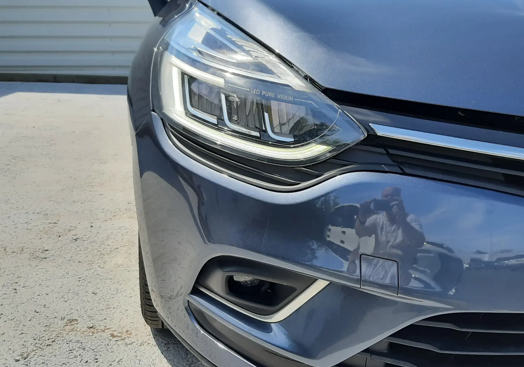 Gros plan sur l'avant gauche d'une Renault Clio IV gris titanium, mettant en valeur le phare LED et la calandre.