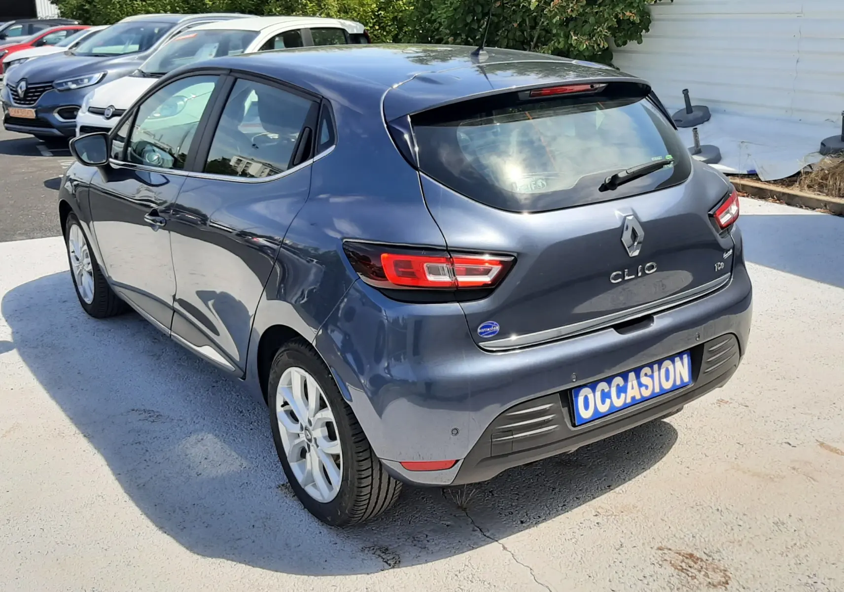 Vue 3/4 arrière droite d'une Renault Clio IV gris titanium avec plaque "OCCASION" sur parking extérieur.