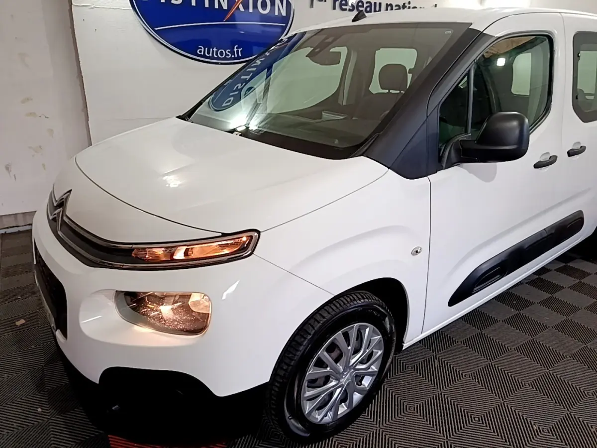 Vue 3/4 avant droit d'un Citroën Berlingo XL blanc avec phares allumés et protections latérales noires.