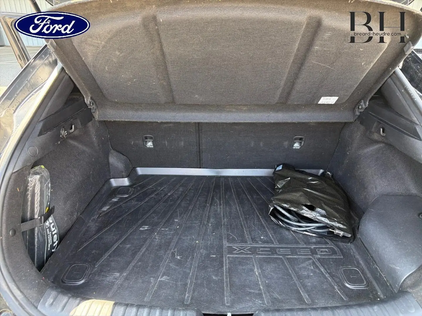 Vue intérieure du coffre ouvert d'un KIA Xceed noir Basalte, avec tapis de coffre marqué Xceed et câble de recharge plié.