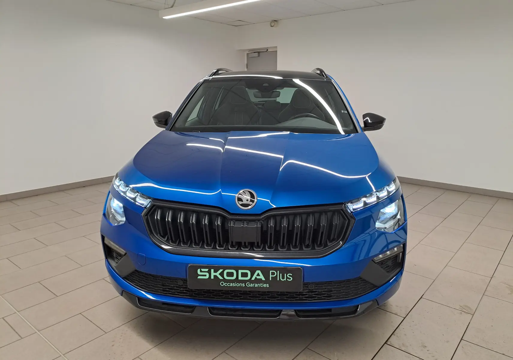 Vue avant d'un Skoda Kamiq 1.5 TSI 150 DSG7 Monte Carlo bleu RS avec calandre noire et feux allumés en intérieur.