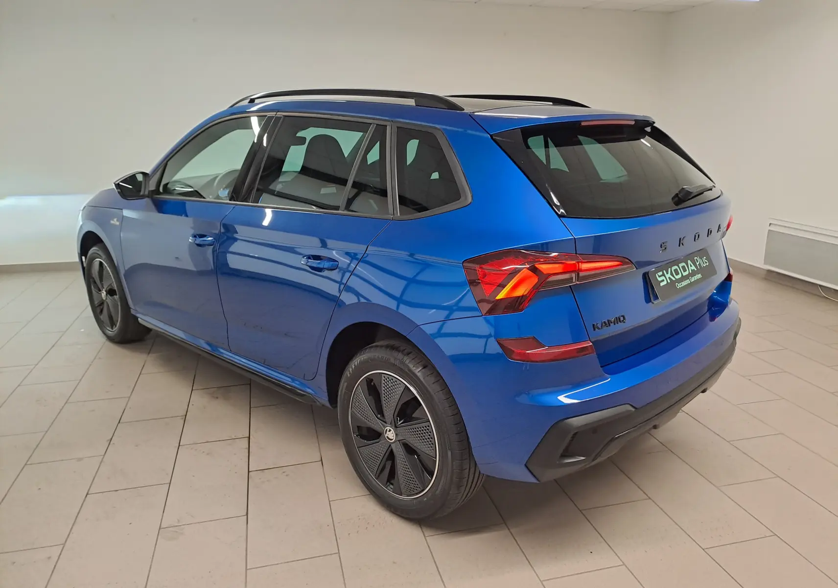 Vue 3/4 arrière droite d’un Skoda Kamiq 2024 bleu RS avec jantes noires et feux arrière allumés en intérieur.