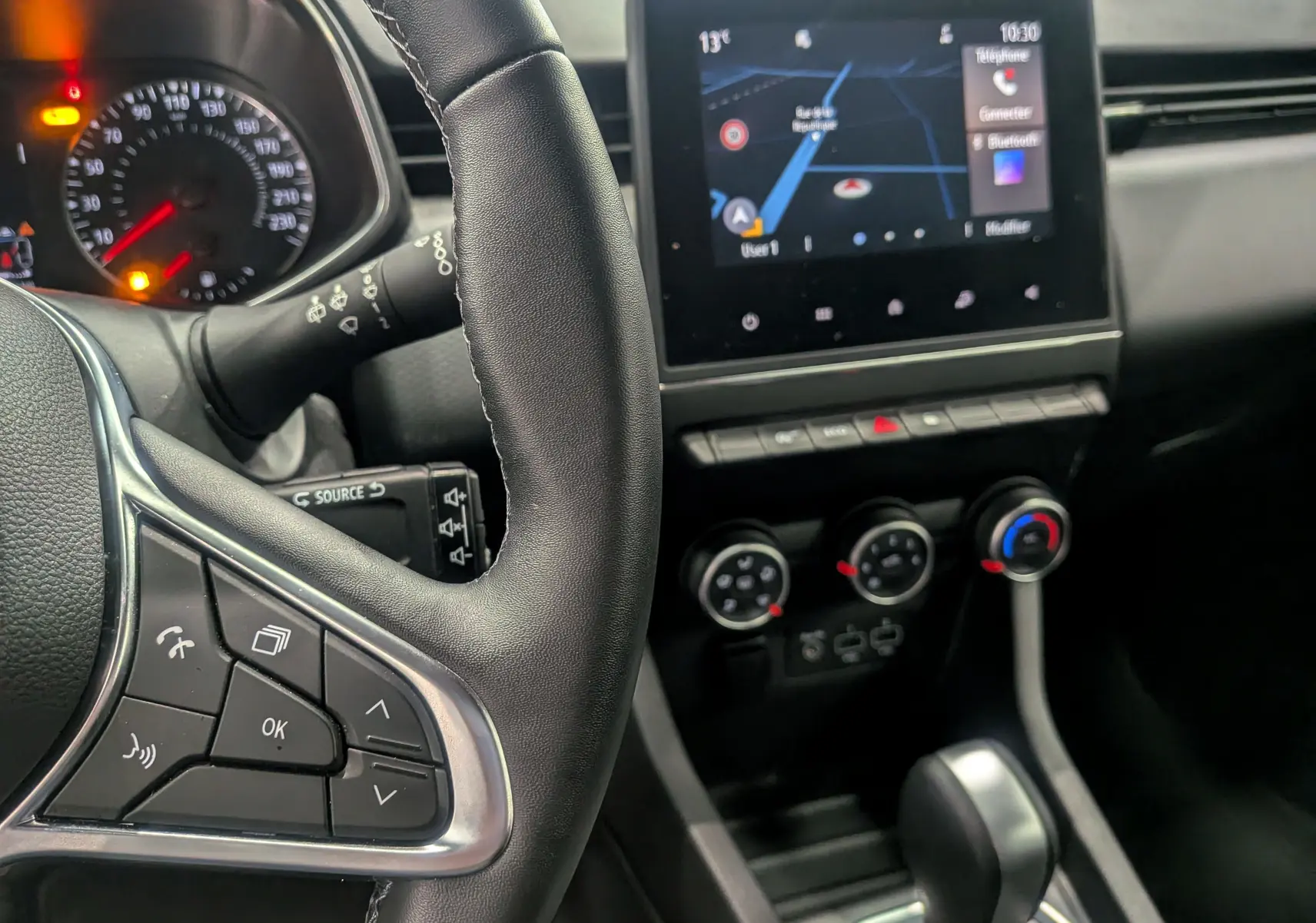 Intérieur de la Renault Clio V blanc glacier, vue rapprochée du volant à gauche et de l’écran tactile central avec navigation.
