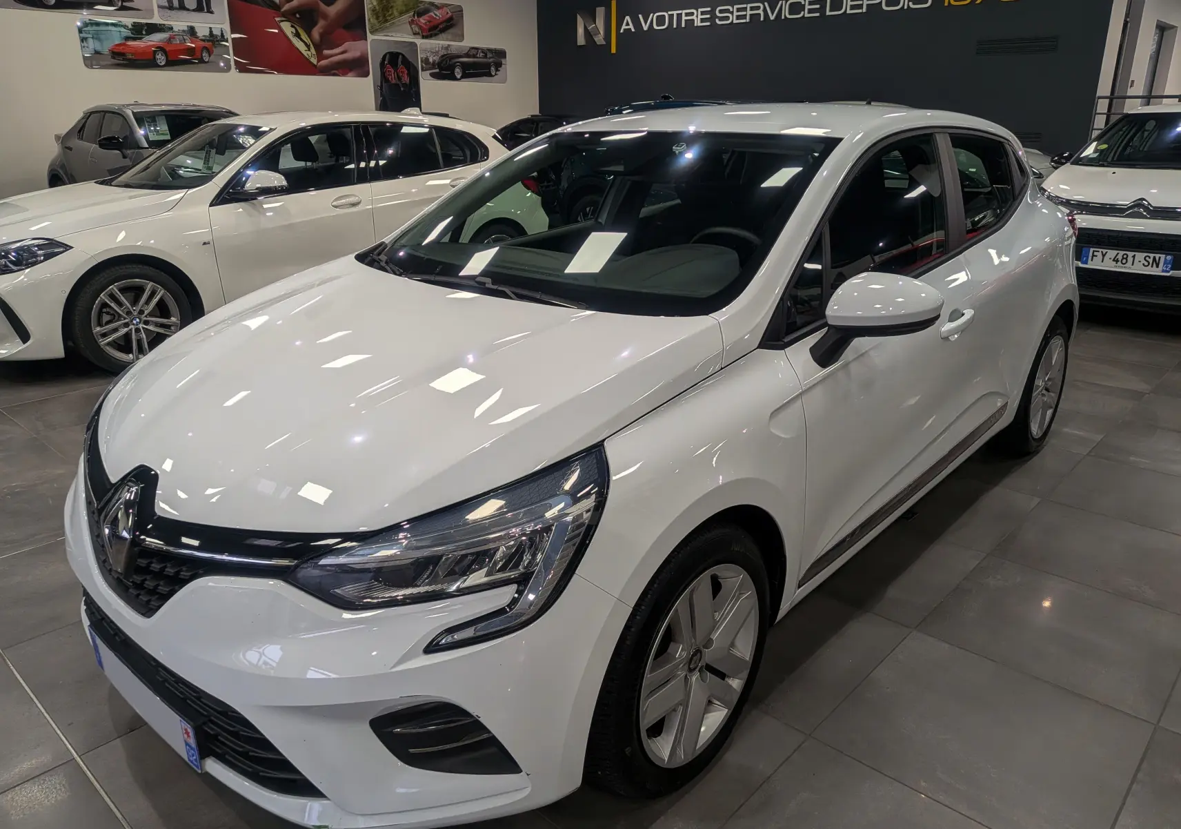 Renault Clio V blanc glacier vue 3/4 avant droit en showroom avec jantes alliage et calandre noire brillante.