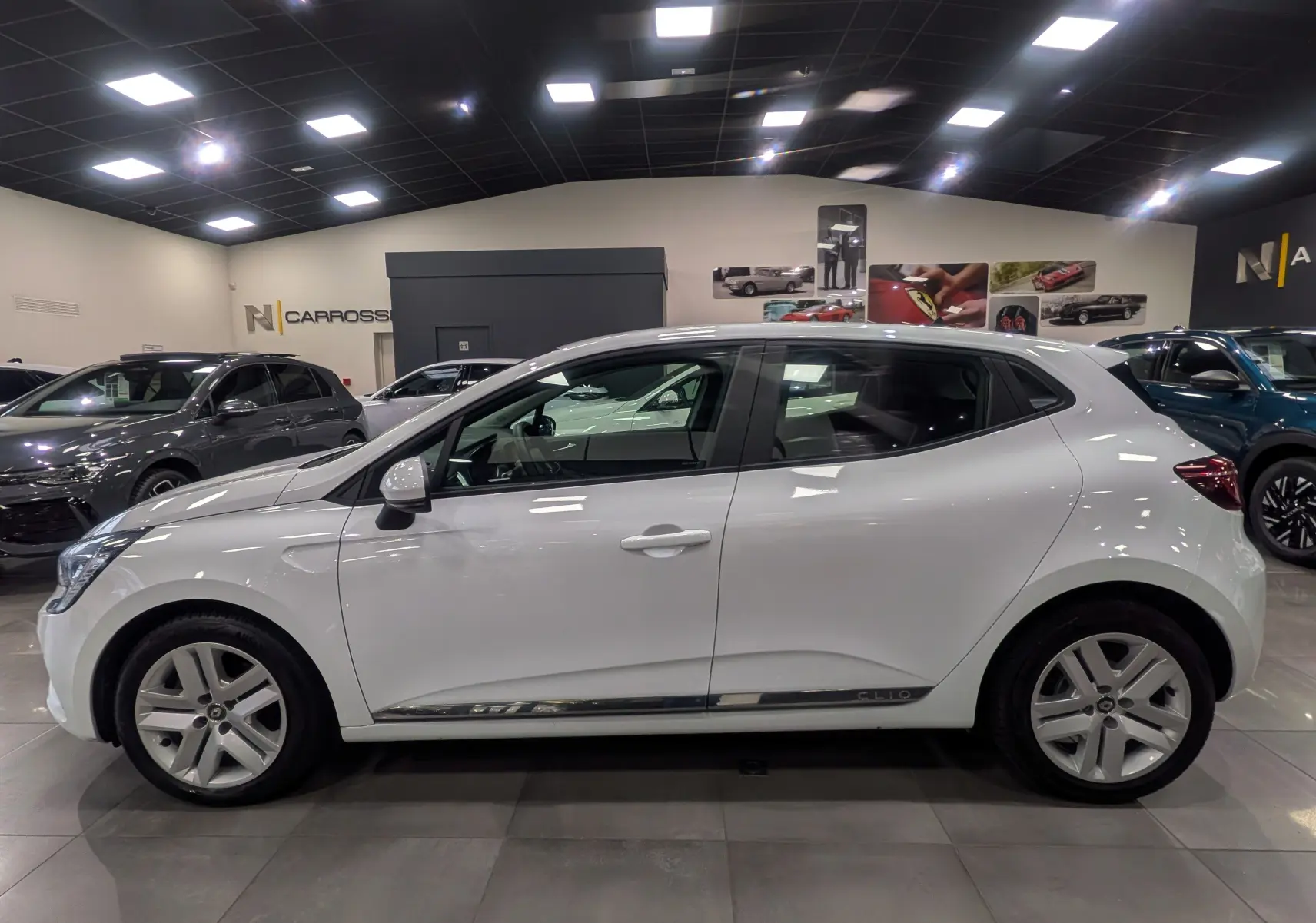 Profil côté gauche de la Renault Clio V blanc glacier, version TCe 100 CH X-TRONIC BUSINESS, en showroom intérieur.