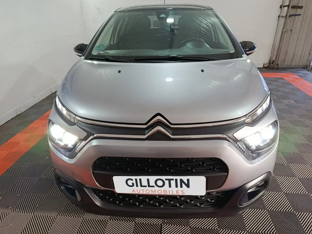 Vue avant d'une Citroën C3 gris foncé avec phares allumés dans un garage au sol carrelé noir et orange.