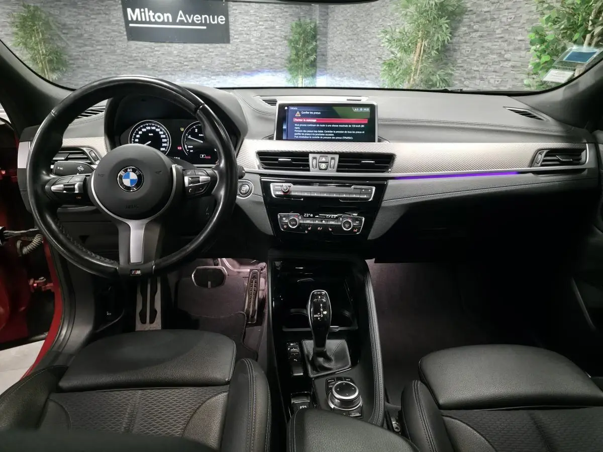 Vue intérieure avant de la BMW X2 orange, tableau de bord avec éclairage d'ambiance violet et volant M cuir noir.