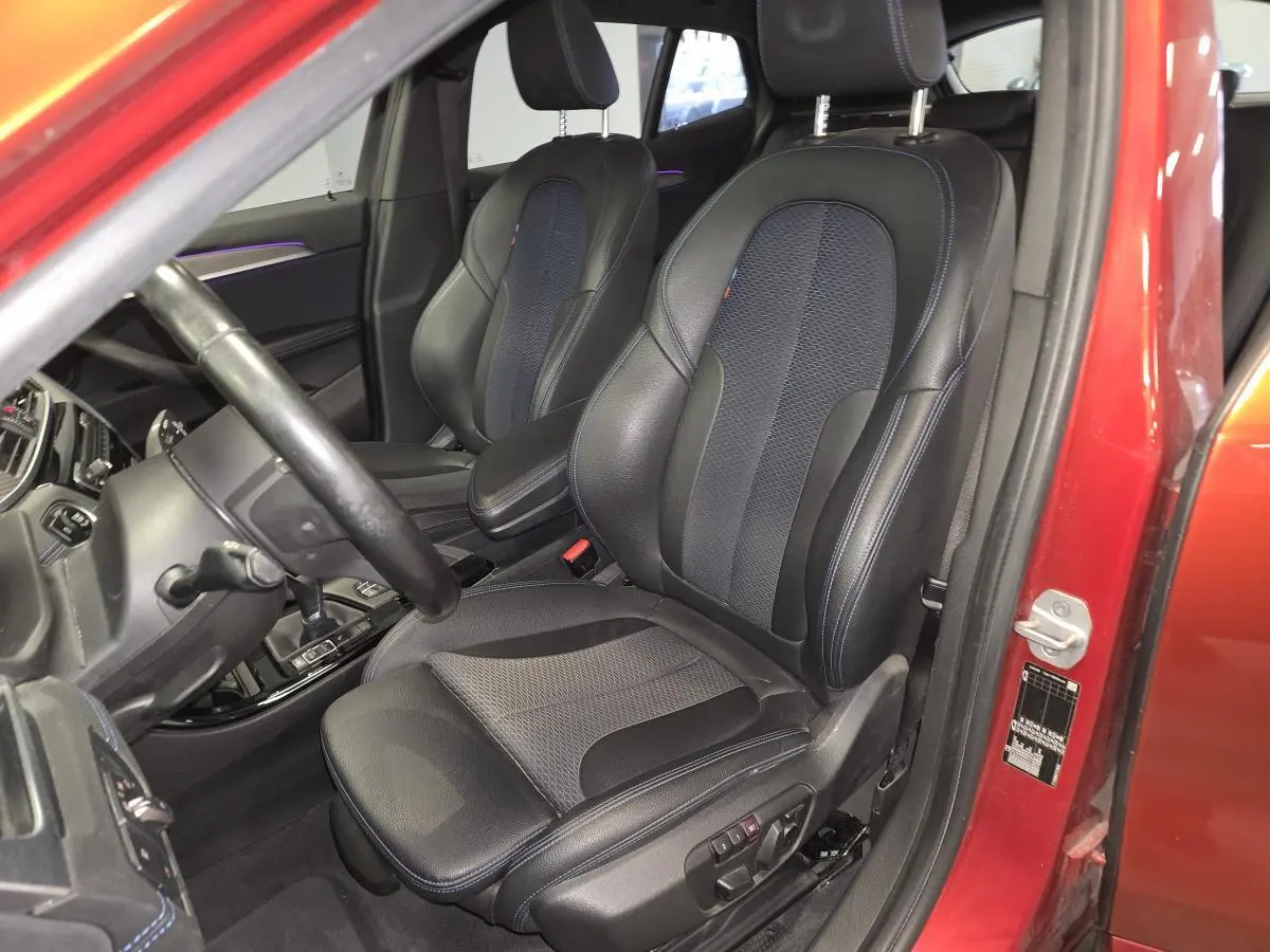 Vue intérieure côté conducteur de la BMW X2 orange 2021, sièges sport en cuir noir avec surpiqûres bleues et éclairage d'ambiance violet.