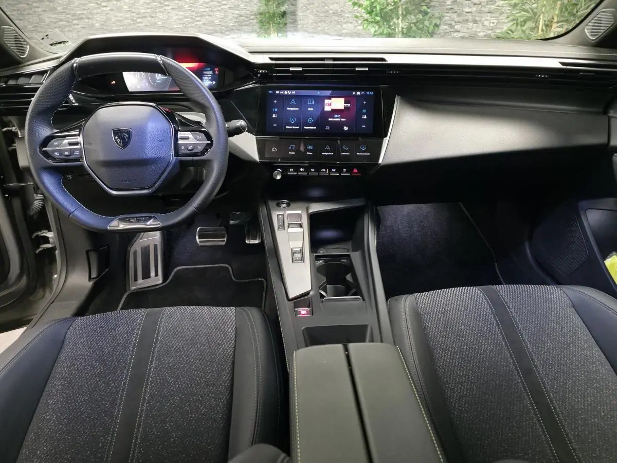 Intérieur Peugeot 408 GT hybride 2025 vu de face, volant cuir perforé, écran tactile central et sièges tissu noir.