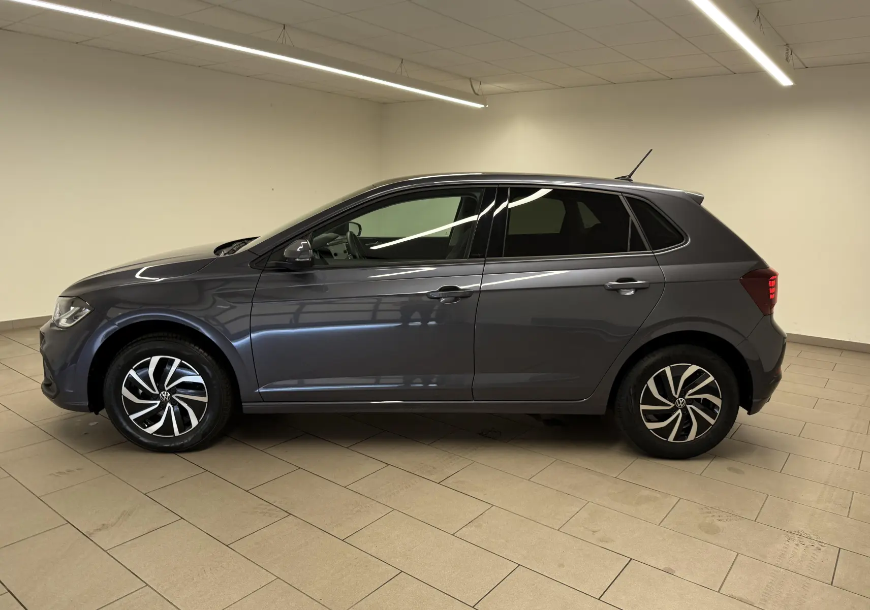 Vue de profil côté gauche d'une Volkswagen Polo gris cendre avec jantes alliage bicolores et vitres teintées, en intérieur.