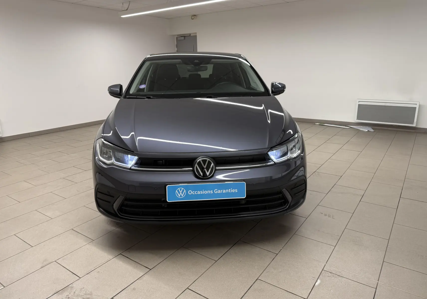 Volkswagen Polo gris cendre vue de face en intérieur avec phares LED allumés et plaque "Occasions Garantis".